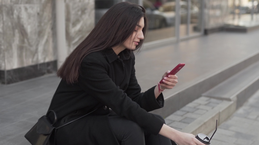 Young charming girl is sitting on a border, drinking a coffee, smiling and clicking on a phone. Girl is wearing a black coat and white keds.