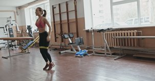Young slender woman doing jumps with resistance band on legs while training in gym - slow motion, raw video record.