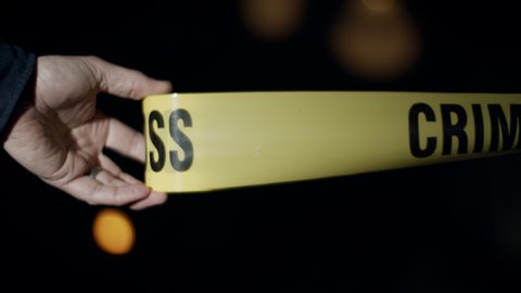 Close-up of a police officer's hand as he rolls out crime scene tape. Flashing emergency lights can be seen in the background.