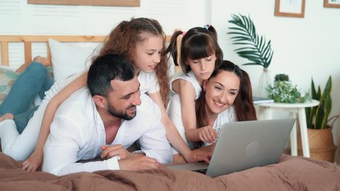 Happy family, parents and two daughters lying on bed in bedroom and play in computer game on laptop, slow motion. Family spends time together at home