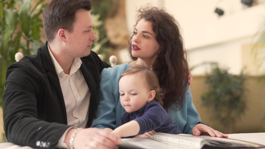 Caucasian Husband and Wife Sitting Together in Restaurant Garden and Talking While Their Baby Girl From Mother's Lap Looking At Menu. Blurred Background