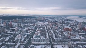 Aerial view of city buildings covered with snow, roads and large river on the background against grey evening sky in winter. Clip. Winter city landscape