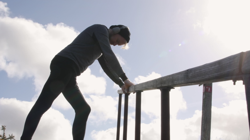 Silhouette shot of a runner stretching on a wooden ramp while listening to music with headphones on a sunny day.