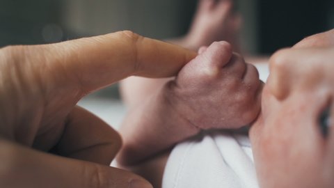 Newborn baby stares at dad while holding his finger. Infant grips the finger of its parent while looking around. Baby in white outfit rests quietly. Tiny fingers of a baby. ZOOM OUT.