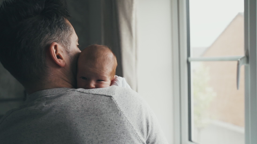 Dad holding baby near a bright window. Father comforts and cuddles infant while standing. Parenthood, fatherhood and love concept. Man with baby against his chest, head on shoulder. Slow motion. Royalty-Free Stock Footage #1031713928