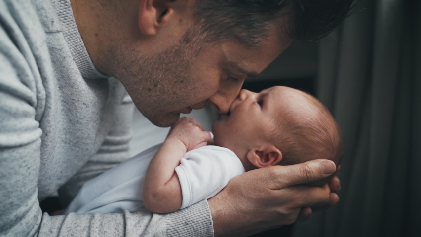 Newborn baby snuggled close to father sucks on dad's nose while looking up at him. Dad holding a tiny infant in his hands. Fatherhood concept with daddy showing affection to baby.