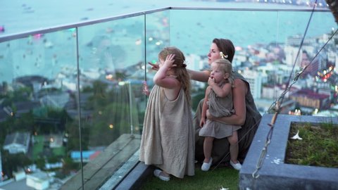 Young mother and her little daughters are looking out the window on evening Pattaya city and coastline. Rooftop viewpoint.