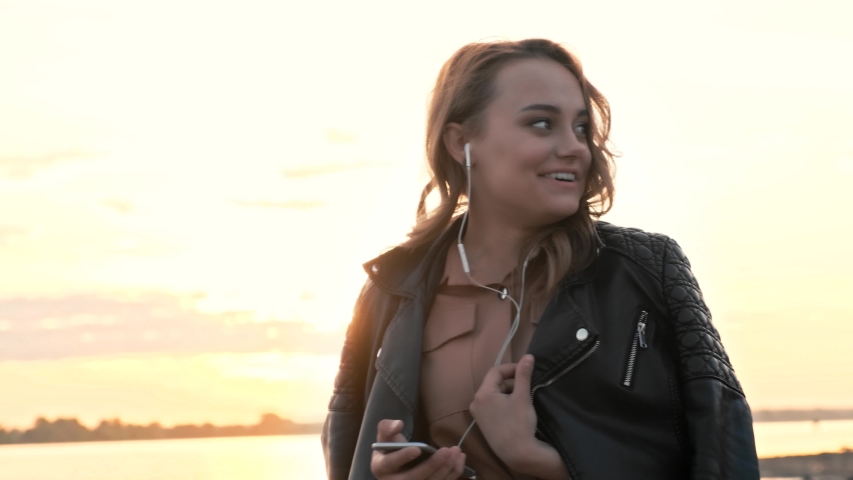 Attractive girl in headphones using smartphone, listening to music and browsing on smartphone. She is looking at the camera and smiling