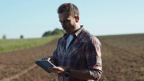 Busy handsome farmer agronomist using mobile technologies studying farm field harvest on sunny day. Young man in shirt holding a tablet looking around working outdoors.
