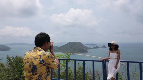 Beautiful young asian tourist woman posing in front of male friend taking photos at seascape viewpoint in Sattahip, Thailand.