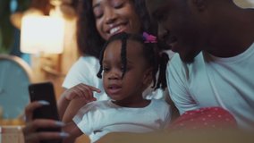 Happy african family with a little cute daughter having fun using smartphone indoors. Portrait of lovely smiling girl lwith braids laughing and watching fun videos with parents on couch.