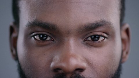 Close-up shot of young black man big eyes blinking and staring at camera. Calm portrait of handsome african guy isolated.