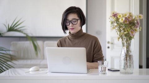 Pan shot of pensive asian woman with short haircut thinking writing a text in laptop. Glass of water on table and a vase with flowers in white interior home office