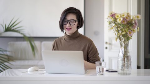 Asian woman in eyeglasses dressed in brown polo neck talking through webcam in white home office. Asian business woman having a video chat through laptop camera