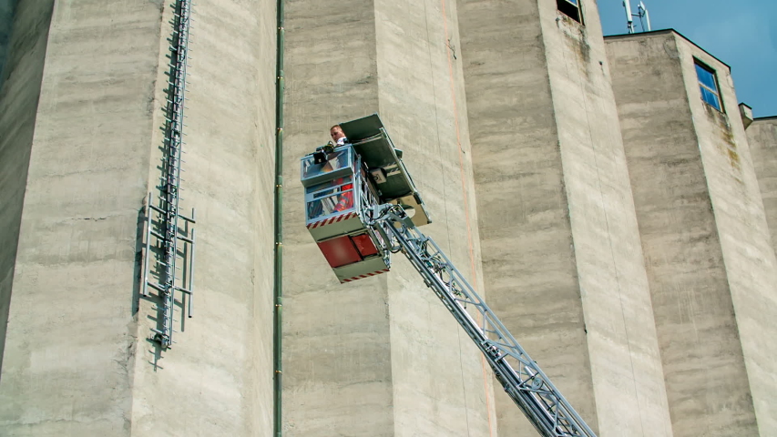 A lift and a ladder are slowly descending. A firefighter is saving a person who got injured when climbing up on the wall of a building.