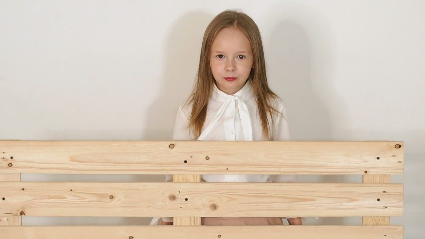 Cute little girl in a white blouse and a long beige skirt posing in the Studio next to a wooden bench on a white background. Portrait of a cute girl of eight years in the Studio. Slow motion.