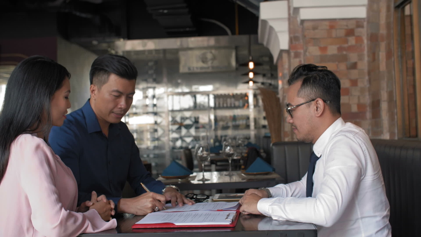 Lockdown of three Asian business partners sitting in restaurant, signing contract and then shaking hands | Shutterstock HD Video #1031756696