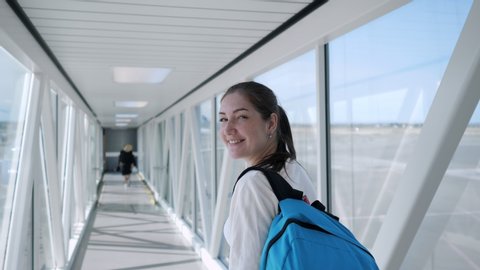 Happy young woman with backpack is going on telescopic gangway to the airplane. She turns around, looks at the camera and smiles.