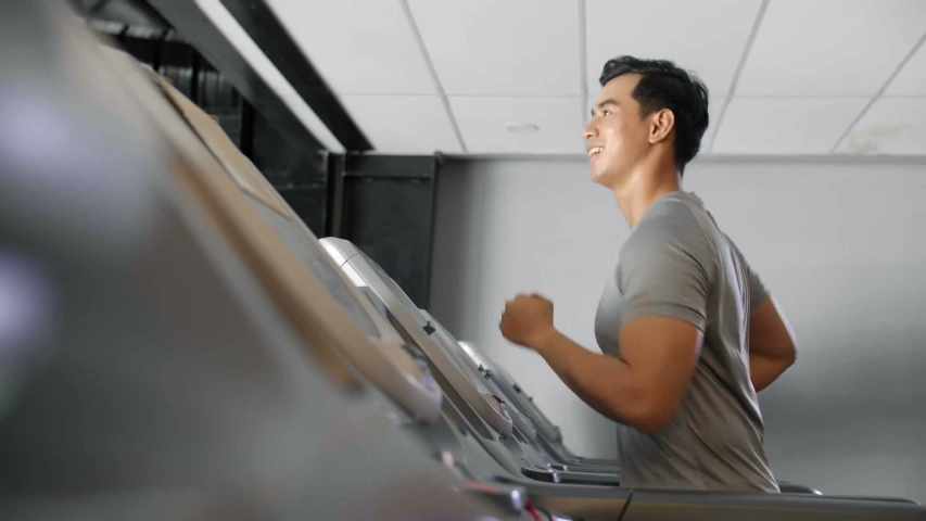 Medium shot of Asian man running on treadmill in gym and smiling