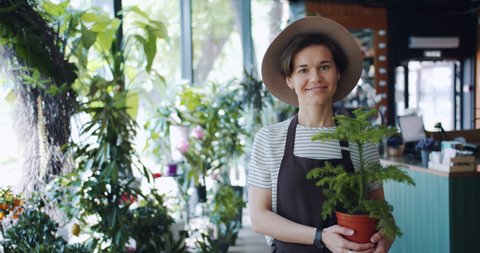 Slow motion portrait of cute young lady in apron and hat holding potted plant in florist's store smiling looking at camera. Business, people and houseplants concept.