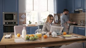 Full shot of glamorous Caucasian businesswoman sitting at kitchen table with laptop, reading newspaper, drinking juice and answering phone call, while husband and toddler son are cooking breakfast - Powered by Shutterstock - Get 15% off with code: PIKWIZARD15