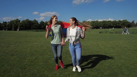 Young joyful lgbt couple running hand in hand across park lawn on windy summer day holding fluttering gay pride symbol. Attractive girlfriends with rainbow flag enjoying freedom of countryside leisure