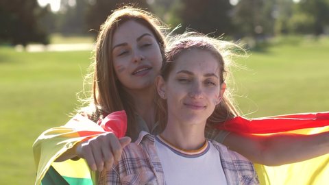 Close-up portrait of happy gay couple wrapped in color lgbt flag and with it painted on cheeks cuddling outdoor. Cheerful pretty girlfriends enjoying embrace and tender kiss spending leisure in park