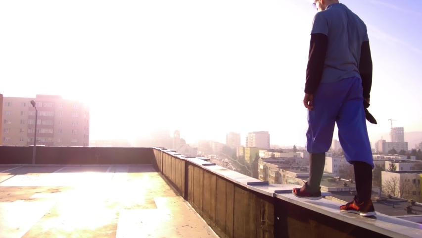 Man walking on the edge of a building rooftop toward bright sunlight in the distance. Buildings of the city far below and on the horizon. TRACKING. - Powered by Shutterstock - Get 15% off with code: PIKWIZARD15