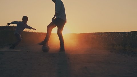 Father and son playing together with ball in football on the field under sunset background. Father and Young Son Playing Soccer.