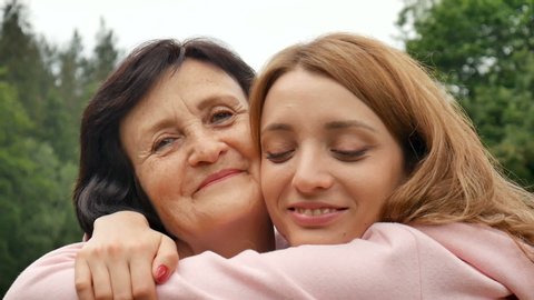 Outdoor close up portrait of smiling happy caucasian senior mother with her adult daughter hugging and looking at the camera. Adult daughter kisses mother, mother's day