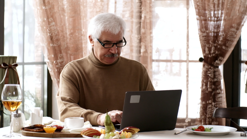 Waist-up shot of 60-something Caucasian gentleman, wearing beige jumper and glasses, sitting at table by large window, having breakfast and drinking coffee, while browsing on laptop