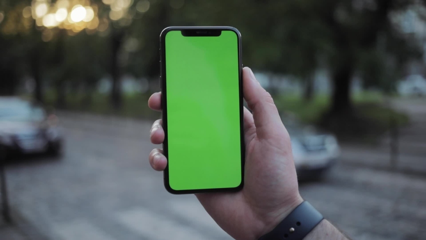 Man using a smartphone mockup screen outside on blurred background. Close-up of male hand holding in vertical position green chromakey smartphone and using touchscreen in the city street. | Shutterstock HD Video #1031820173