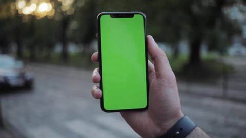 Close-up of sociable man checking news using a smartphone outdoors. Man's hand with smartphone greenscreen mockup against busy street in the city.