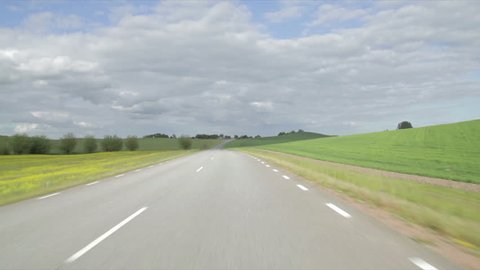 Driving a car up a hill on a tarmac road, Mounted camera, front view. Country road with fields and trees along the way,  cloudy sky, day. slow Speed