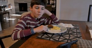 Young boy 8 - 10 years old eating a sausage patty for breakfast while watching a mobile phone sitting alone at a dining room or kitchen table in a house during the day