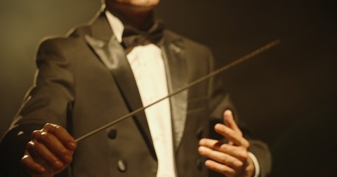Male orchestra conductor wearing tux standing in front of music stand, controlling musicians by moving his hands and baton. Studio shot on black background 4k footage