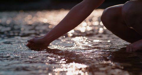 slow motion of a woman arm playing with water on a river during the sunset, woman hand in the water with a backlight 