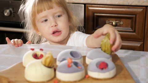 Portrat of little cute girl is decorating small cake with raspberry in kitchen table at home and she is eating berries.
