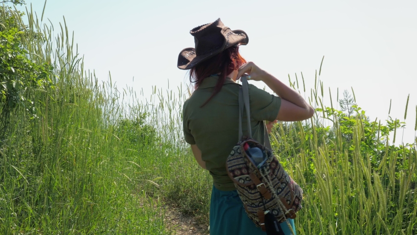 A young red-haired traveler girl in a cowboy hat with a backpack walks along a mountain path overgrown with grass and shrubs.
