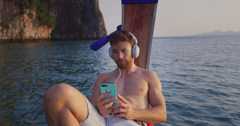 Man listening to music on smartphone with headset on long tail boat tour in Asia Thailand, with lime stone cliffs in South East Asia at sunset. Slow motion hand held. Shot on red.