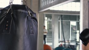 Powerful shirtless male punching bag fiercely while boxing on blurred background of modern gym