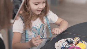 Close up video of beautiful little girl painting ceramic eggs. She checks her work, soaks brush into cup of water to get new gouache colour. While doing that, her mother takes photos of whole process