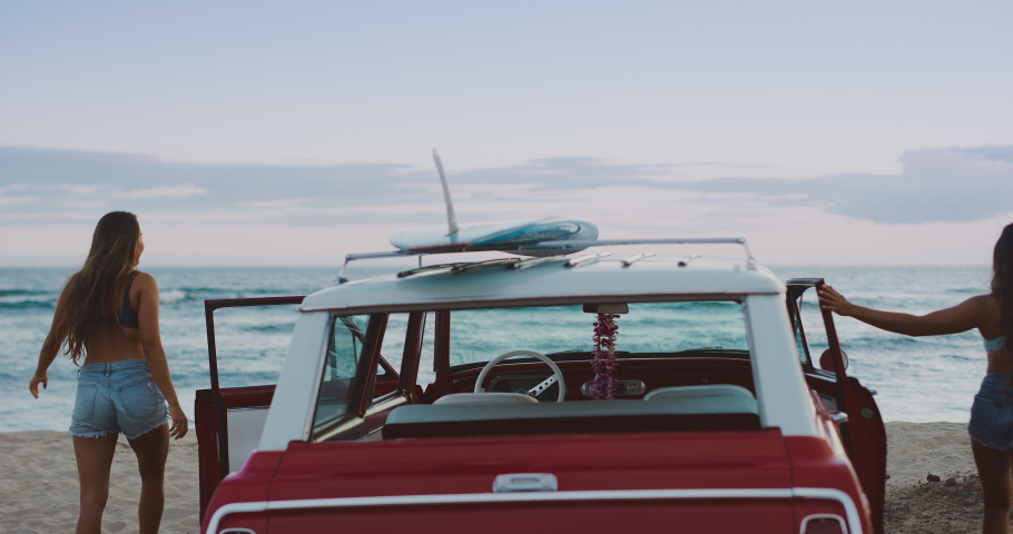 Two friends arriving at the beach in a vintage beach cruiser car to go surfing, two girls getting out of a vintage car at the beach and raising their arms in joy, hawaiian island surf adventure