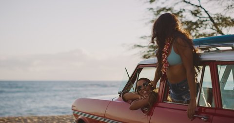 Two young attractive women in a vintage beach cruiser smiling and looking out at sunset, happy girls raising arms in celebration and smiling, friends at the beach, hawaiian island surf adventure
