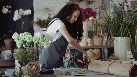 Attractive women with dark curly hair is smiling while making a bouquet of dried flowers and cutting off excess branches with a pruner at flower shop.