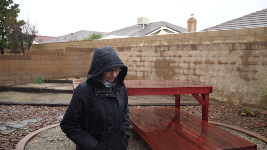 An elderly woman walking in a raincoat and hood during a winter rain storm as drops fall under a cloudy sky in slow motion.