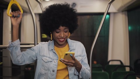 Smiling young african american woman holds the handrail and watching video on the smartphone at public transport. Night time. Close-up. City lights background.