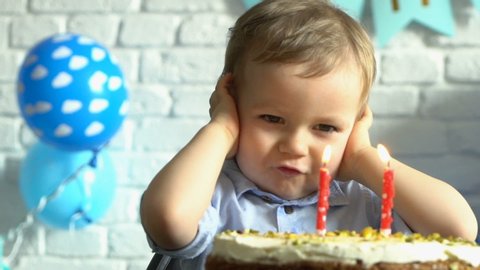 Little baby boy celebrating his second birthday and blowing candle on homemade baked cake indoor, slow motion