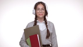 Cheerful young brunette girl student with books listening music with wireless headphones and dancing while looking at the camera over white background isolated