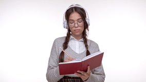Concentrated young brunette girl student smiling and making notes in notebook while listening an audio book with wireless headphones over white background isolated